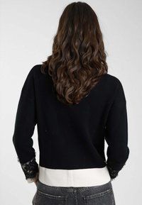 Maglione nero a maniche lunghe, con cintura bianca e dettagli in pizzo sui polsini. Tessuto morbido, vestibilità comoda e design semplice.