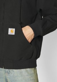 Schwarzer Kapuzenpullover mit Reißverschluss aus Baumwolle, mit gerippten Bündchen und Saum. Verfügt über eine Fronttasche und einen quadratischen Logoaufnäher auf der linken Seite.