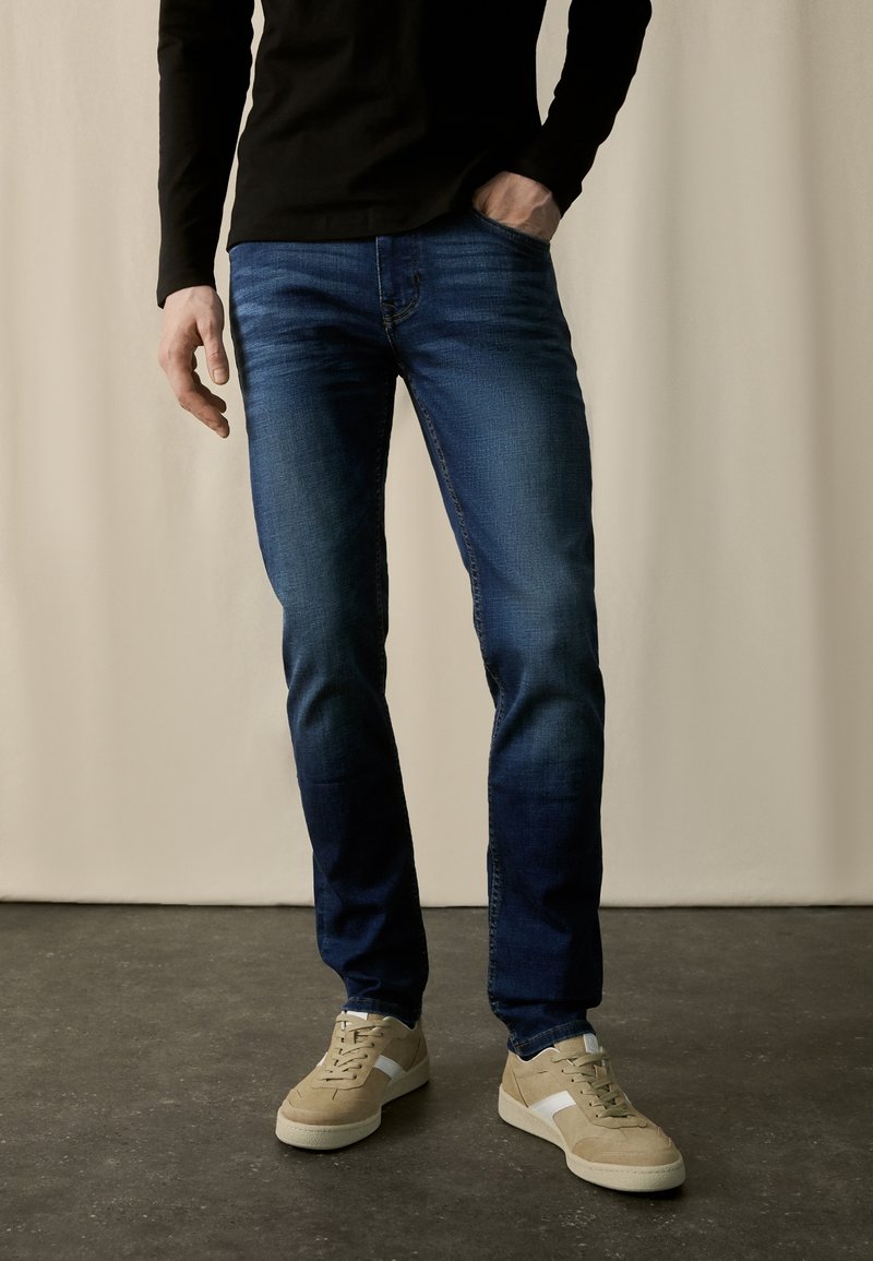 Homme portant un jean slim bleu foncé, une chemise noire à manches longues et des baskets beiges, debout sur un sol sombre avec un fond clair.