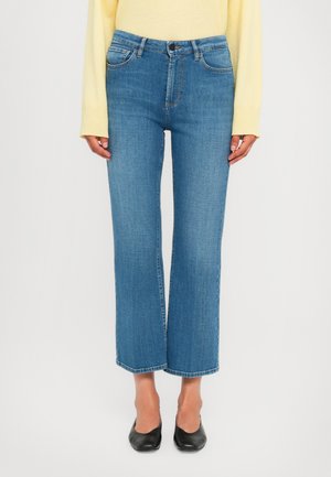Woman wearing cropped blue jeans, yellow long-sleeve top, and black pointed-toe flats standing against white background.