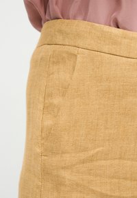 Κοντινό πλάνο της μέσης μπεζ λινής φούστας με πλαϊνή τσέπη, φορεμένη με πουκάμισο μωβ μωβ που είναι μπουκωμένο μέσα.