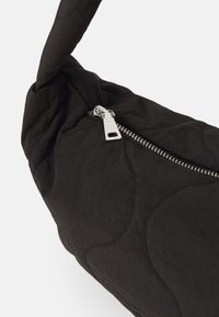 Torba od crnog kviltanog materijala s zakrivljenim oblikom, koja sadrži detalj s patent-zatvaračem i jedan ručni remen. Glatka tekstura i minimalistički dizajn.