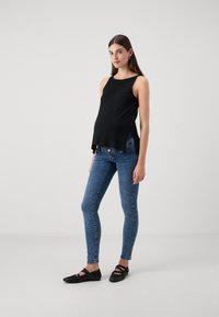 Top de maternidad negro sin mangas con aberturas laterales, combinado con jeans skinnies azules y bailarinas negras con tiras entrecruzadas.
