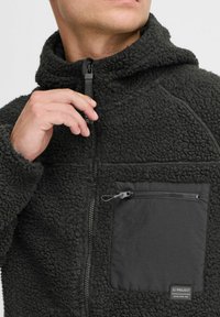 Veste en polaire noire avec une texture douce et duveteuse, dotée d'une poche poitrine zippée en tissu lisse. Elle présente un col haut et une fermeture zippée.