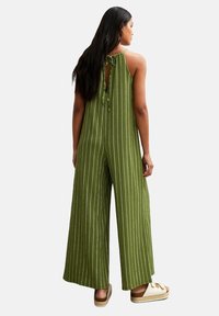 Tuta verde a righe con legamenti sul retro, gambe larghe, tessuto testurizzato e apertura a chiave. Abbinato a sandali con plateau di colore chiaro.