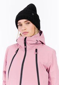 Roze waterdichte jas met hoge kraag, zwarte ritsen en verstelbare koorden, gecombineerd met een zwarte geribbelde gebreide muts.