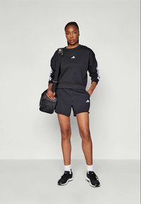 Juodas sportinis kostiumas, sudarytas iš viršaus su ilgomis rankovėmis ir šortų, abu su balta Adidas logotipu. Rinkinį papildo juoda petinė rankinė ir sportiniai batai. Paprastas pilkas fonas.