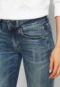 Blå denimjeans har en klassisk design med fem fickor, subtil blekning och en metallknappstängning, med synliga sömmar längs sömmarna.