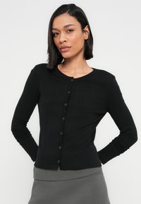 Μια γυναίκα με ίσια σκούρα μαλλιά φοράει μια εφαρμοστή μαύρη ζακέτα με κουμπιά και μια γκρι ριμπ πλισέ φούστα, στέκεται με τα χέρια πίσω από την πλάτη της.