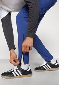 Calças atléticas azuis com fechos de tornozelo com zíper, combinadas com tênis preto e branco. A parte superior tem mangas longas cinzentas e corpo branco.