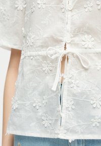 Blouse blanche brodée de fleurs avec manches courtes, fermeture par boutons et lien, portée sur un jean denim bleu clair.