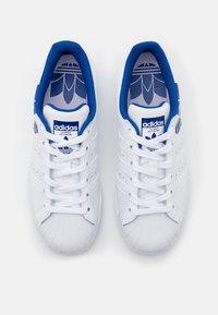 Valkoiset nahkakengät sinisillä yksityiskohdilla, joissa on kuminen varvaskuppi, teksturoitu päällinen ja kuusi nauhoitusrei'it. Adidas-logo kielessä.