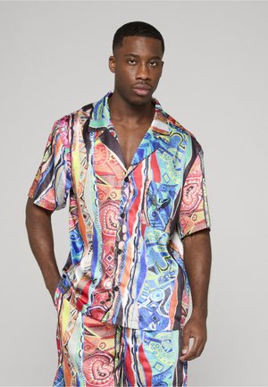 Hombre vestido con una camisa de manga corta con botones y un patrón colorido, combinada con pantalones cortos a juego, de pie contra un fondo liso de color gris claro.