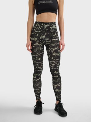 Kvinde iført sort sports-bh og højttaljede leggings med camouflage-mønster samt sorte sneakers, stående foran en ensfarvet baggrund.