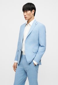 Costume bleu clair en tissu texturé, comprenant une veste à simple boutonnage, des revers crantés et un pantalon sur mesure ; chemise blanche en dessous.