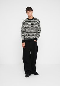 Μαύρος και λευκός πουλόβερ με σχέδιο, με στρογγυλή λαιμόκοψη, μακριά μανίκια και ριμπ μανσέτες, σε συνδυασμό με φαρδιά μαύρα παντελόνια και παπούτσια.