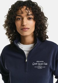 Navyblauwe ritsvest gemaakt van katoen, met op de borst witte geborduurde tekst "New Haven Gant Sports Club 1949 USA".