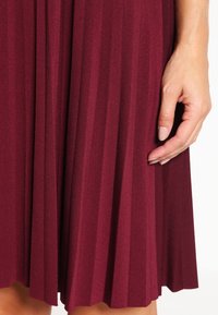 Burgundy plisserad klänning i mjukt tyg, med smala plisseringar och en slät textur. En hand som vilar på tyget framhäver designens detaljer.