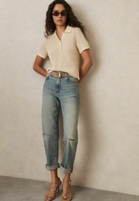 Polo shirt a coste beige con maniche corte, abbinato a jeans in denim chiaro. Una cintura marrone e sandali con tacco alto color beige completano l'outfit.