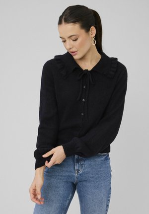 Femme ajustant la manche d'un cardigan noir à boutons avec col volanté, portant des boucles d'oreilles créoles en or et un jean bleu sur un fond uni.
