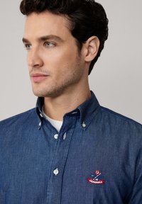Donkerblauwe denim overhemd met witte button-down kraag, voorzien van een rode geborduurde logo op de borst. Soepele textuur.
