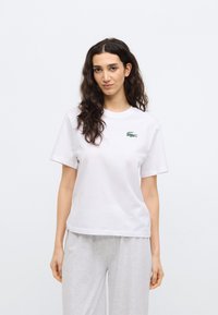Camiseta blanca de algodón con mangas cortas, que presenta un logo de cocodrilo verde bordado en el área del pecho izquierda. Corte plano y relajado.