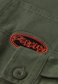Green fabric with a textured finish features a black oval patch embroidered with the word "FORMER" in orange lettering and a button detail.