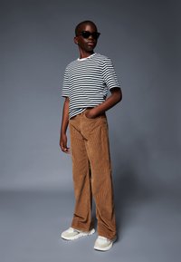 Pruhované tričko v čierno-bielej kombinácii, hnedé kordové nohavice a šedé tenisky. Slnečné okuliare dodávajú outfitu moderný nádych.