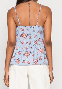Top peplum floral bleu à fines bretelles, avec des motifs de roses roses et blanches, en tissu léger et à coupe fluide.