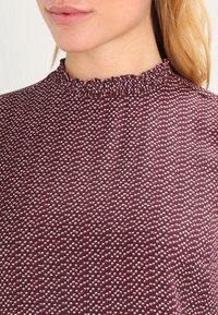 Burgundiblus med volangkrage, med ett strukturerat mönster av små vita prickar. Slät tyg med en kroppsnära design.