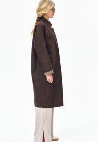 Kobieta z blond warkoczem, ubrana w oversize'owy ciemnobrązowy płaszcz, jasnobeżowe szerokie spodnie, białe buty i okulary, stojąca bokiem.