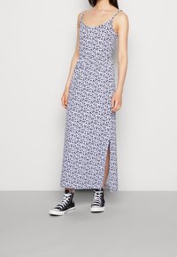 Robe longue sans manches à motif floral, de couleur violet clair avec des accents noirs ; elle comprend une ceinture à la taille et une fente latérale, portée avec des baskets montantes noires.
