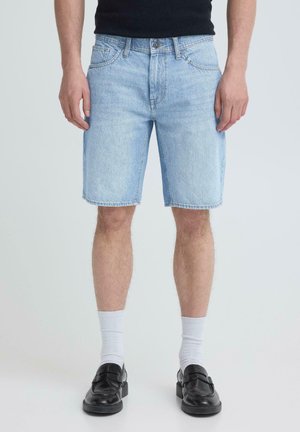 Mężczyzna ubrany w jasnoniebieskie dżinsowe szorty, białe skarpety sięgające do kostek oraz czarne skórzane mokasyny, stojący na tle jednolitego jasnego tła.