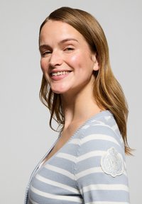 Ljusblå och vit randig cardigan i mjukt stickat material, med en rund emblemdetalj på vänster axel. V-ringad design.