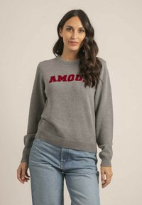 Pull gris avec un col et des poignets côtelés, arborant le texte brodé rouge "AMOUR" sur la poitrine, porté avec un jean bleu clair.