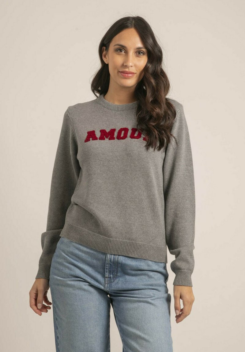 Pull gris avec un col et des poignets côtelés, arborant le texte brodé rouge "AMOUR" sur la poitrine, porté avec un jean bleu clair.