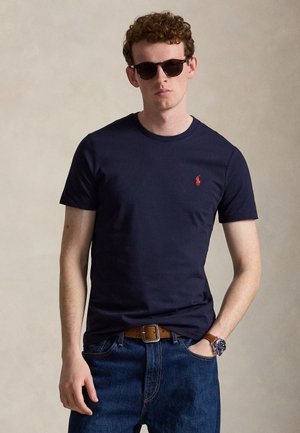 Giovane uomo con capelli ricci che indossa occhiali da sole scuri, maglietta blu navy, jeans blu, cintura marrone e orologio da polso, in piedi davanti a uno sfondo semplice.