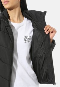 Personne portant un t-shirt blanc avec le logo Vans "Off The Wall" et tenant ouverte une veste noire zippée pour montrer la doublure intérieure.