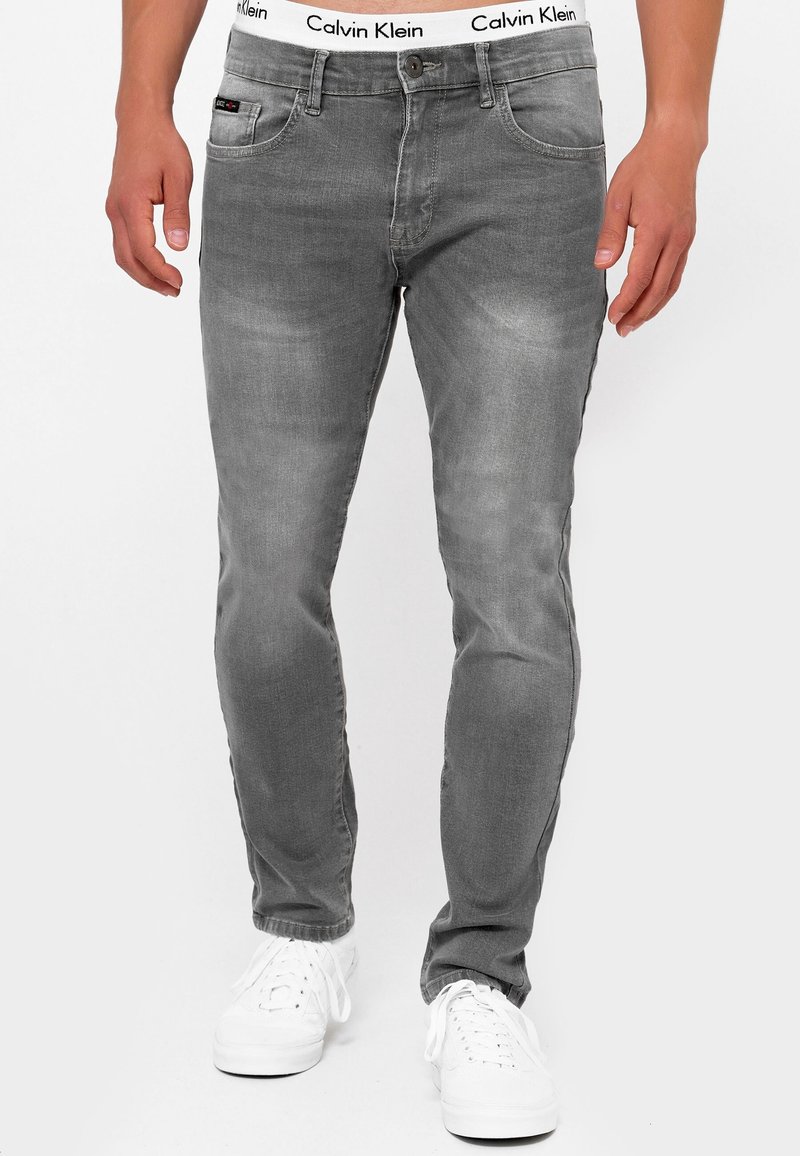 Homme portant un jean gris ajusté avec un sous-vêtement blanc à ceinture Calvin Klein et des baskets blanches, debout devant un fond blanc.