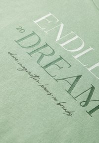 Světle zelené bavlněné tričko s velkým bílým a tmavě zeleným textem. Design zahrnuje frázi "NĚKONEČNÝ SEN" s menším textem níže.