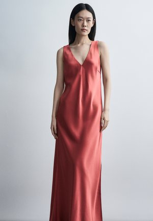 Hosszú fekete hajú nő korallszínű, ujjatlan, mély V-nyakú szatén ruhában áll egyszerű világosszürke háttér előtt.