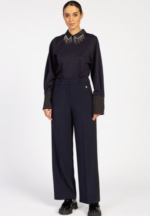 Vrouw die staat in een zwart longsleeve shirt met een versierde kraag, en een brede navy broek met zwarte zijpanelen en zwarte platform schoenen.
