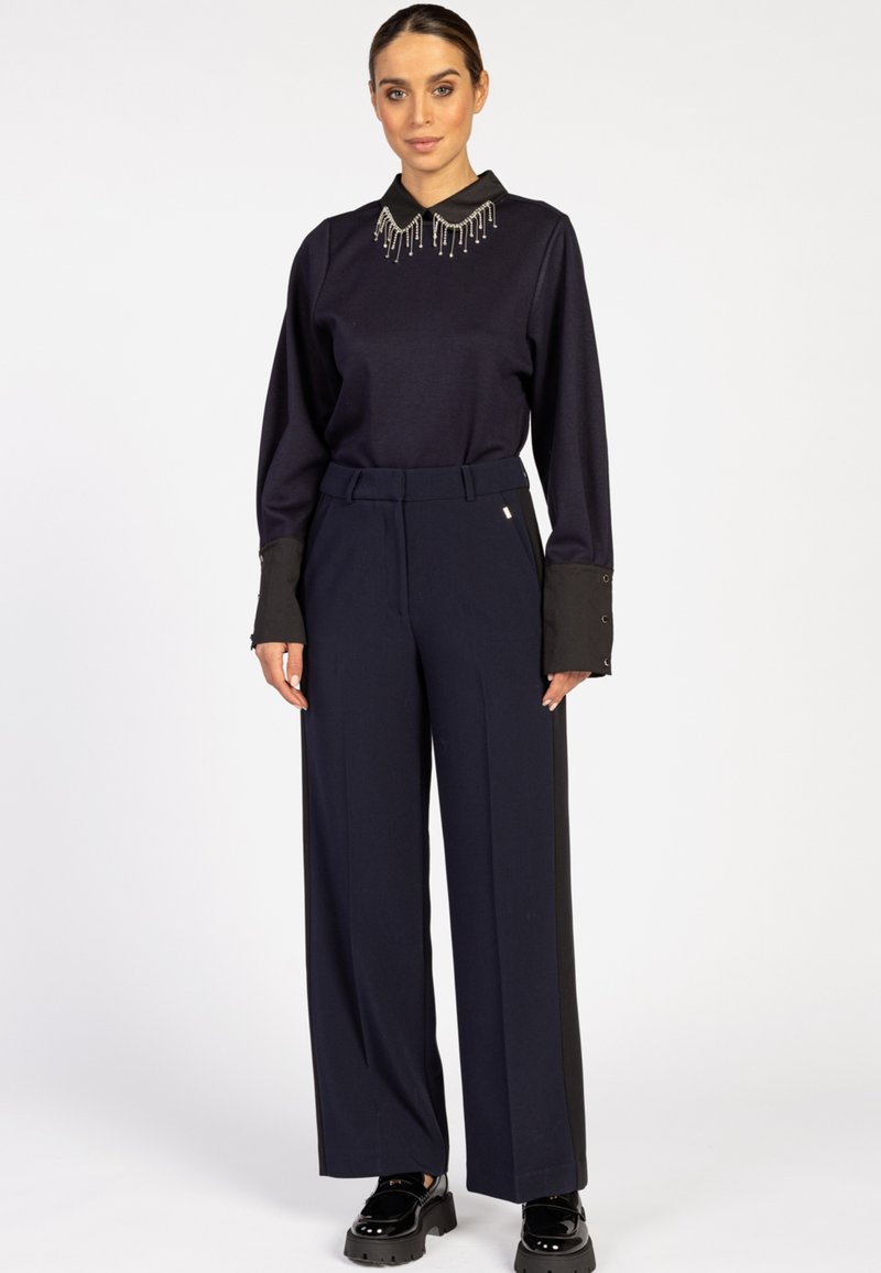 Vrouw die staat in een zwart longsleeve shirt met een versierde kraag, en een brede navy broek met zwarte zijpanelen en zwarte platform schoenen.