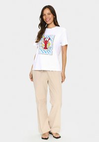 T-shirt blanc présentant un graphique de homard en rouge et jaune, sur un fond à carreaux bleus. Associé à un pantalon large beige.