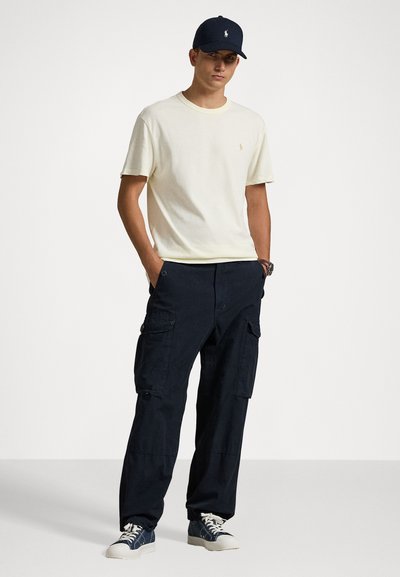 T-shirt de algodão amarelo claro, calças cargo azul-marinho com bolsos e boné de basebol azul-marinho. Ténis casuais completam o visual.