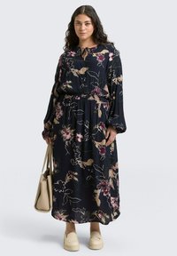 Bloemen maxi-jurk in marineblauw met lange mouwen en een elastische taille. Bevat een striksluiting bij de hals en een persoon die een beige tas vasthoudt.