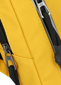 Gelber Stoff-Rucksack mit strukturierten Oberflächen, ausgestattet mit schwarzen Reißverschlüssen und Nähten sowie metallischen Reißverschluss-Ziehern mit detailliertem Design.