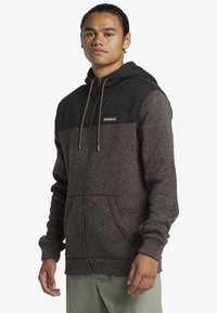 Brown zip-up hoodie with a black upper section, featuring a drawstring hood and subtle texture. Includes side pockets and brand patch detail.