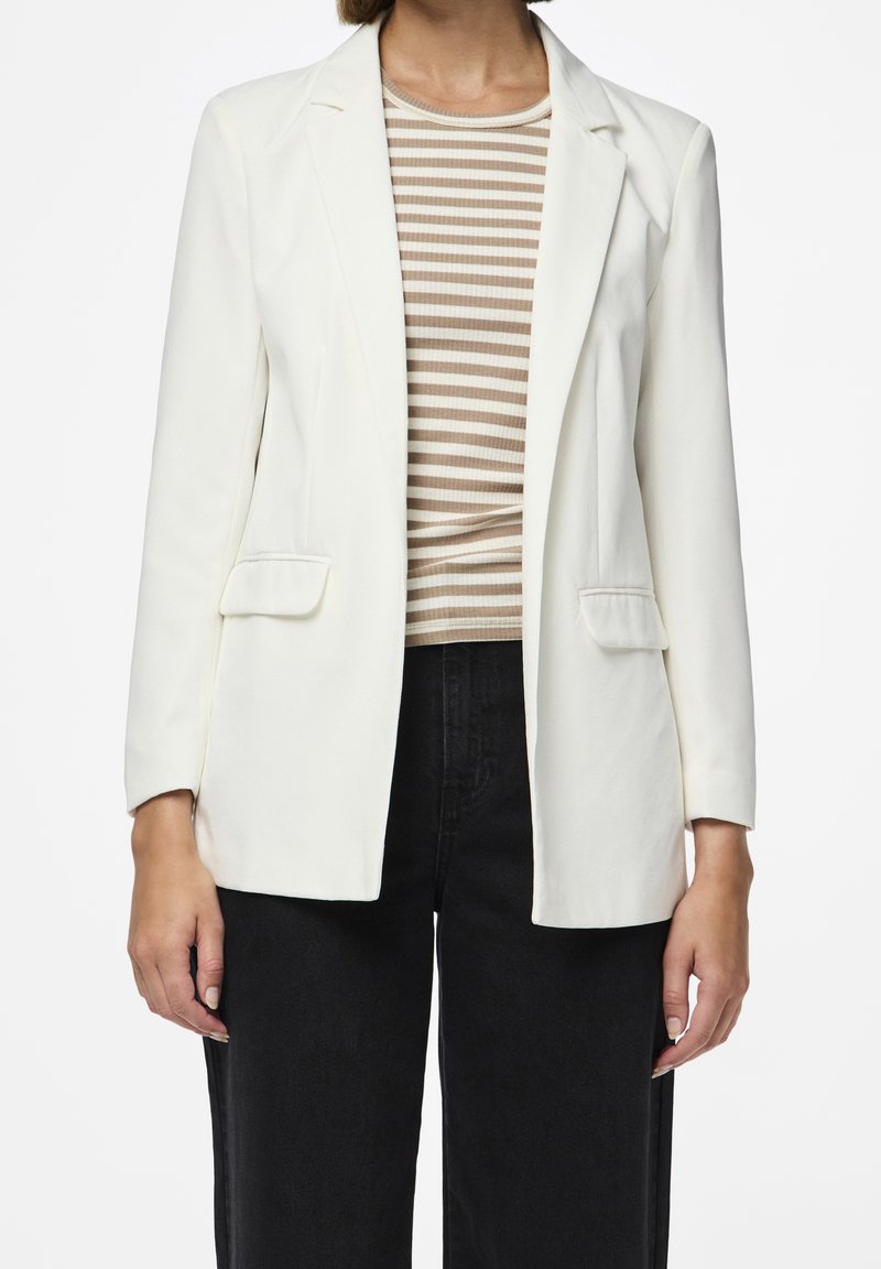 Femme portant un blazer blanc par-dessus une chemise beige et blanche rayée et un pantalon noir, debout devant un fond blanc uni.