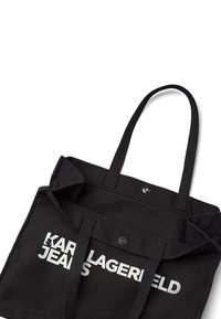 Borsa a spalla nera in canvas con due manici robusti, dotata di una chiusura con bottone a pressione argentato e con il testo bianco in grassetto "KARL LAGERFELD JEANS."